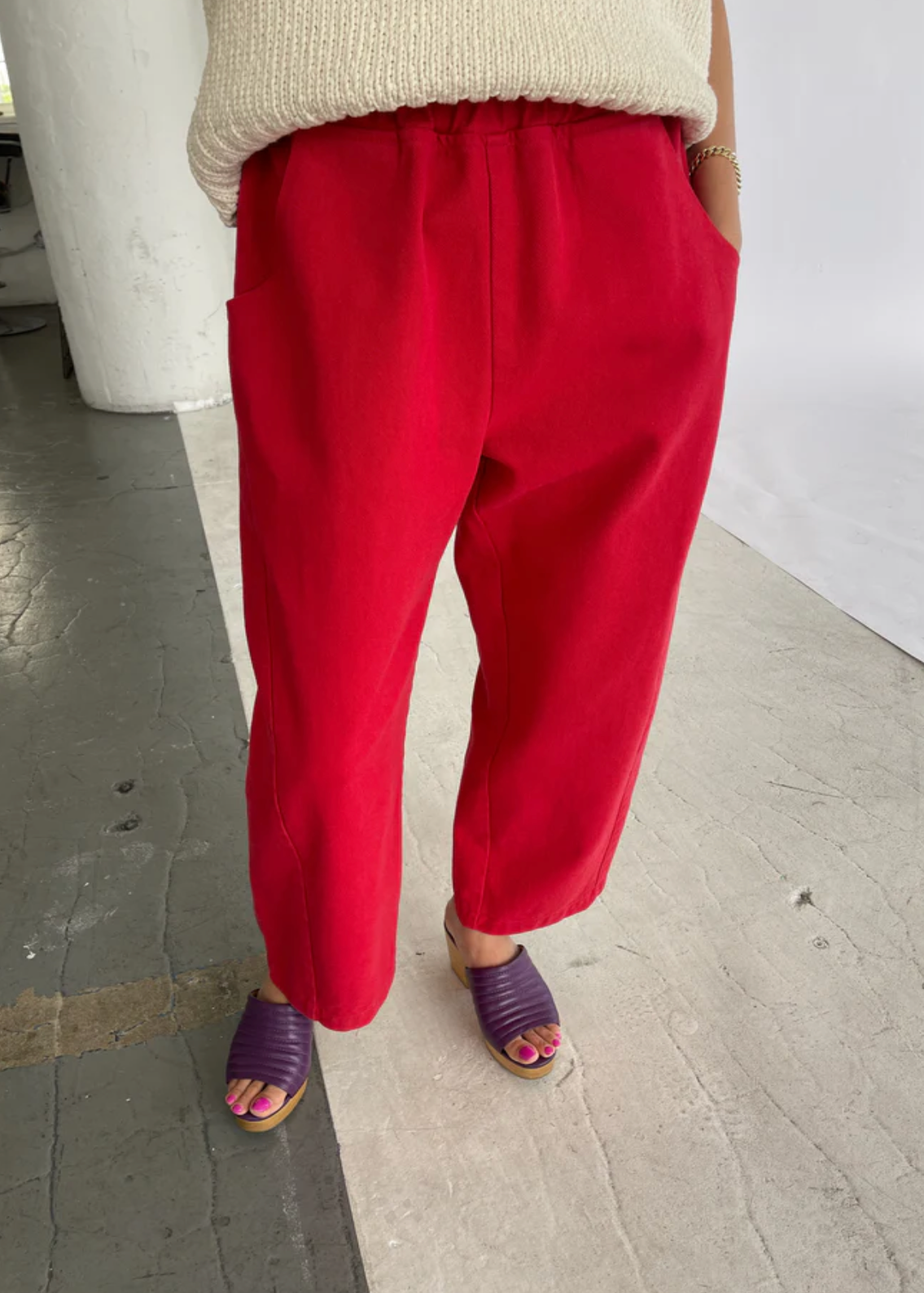 A person wearing bright red cropped pants with a cream knit top and purple wedge sandals, standing on a concrete floor in an indoor space.