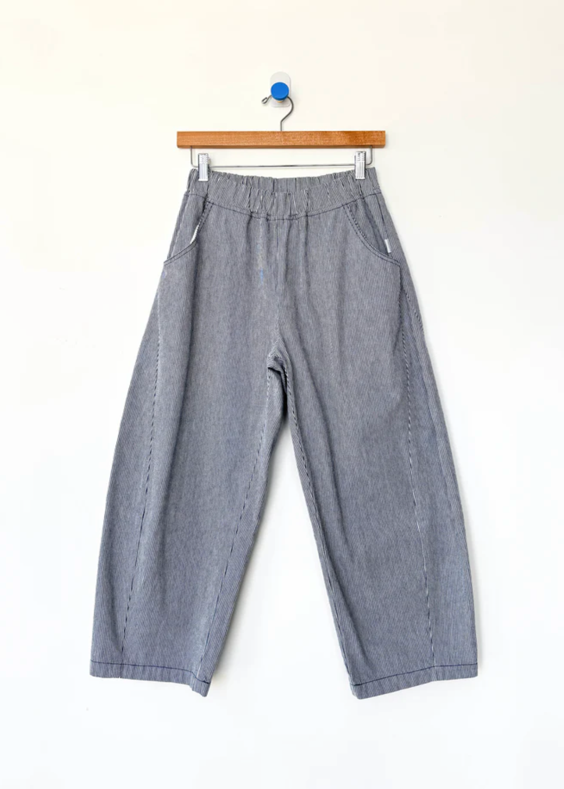 A pair of loose, wide-leg striped pants with a relaxed straight-to-slightly-cropped leg, hanging from a wooden hanger against a plain white wall.