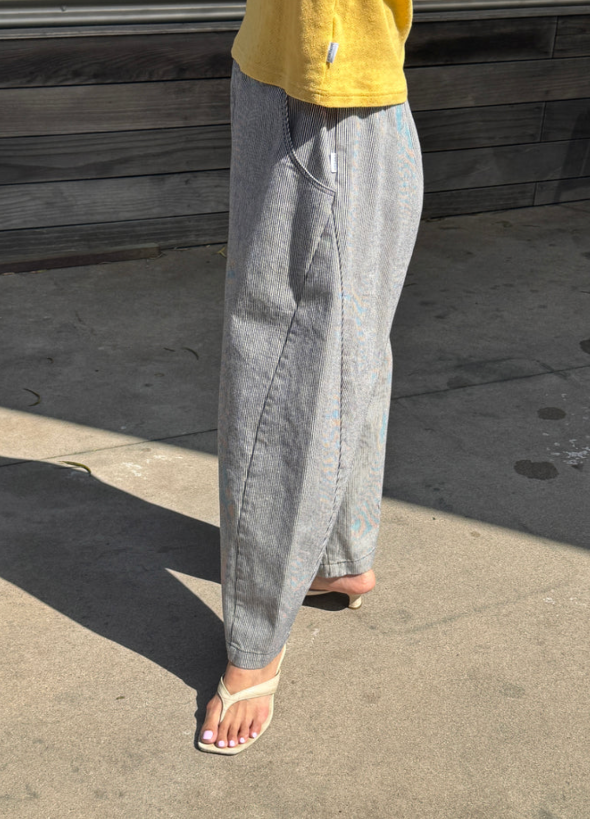 Person wearing striped pants and a yellow top standing on a concrete surface with wooden planks in the background.
