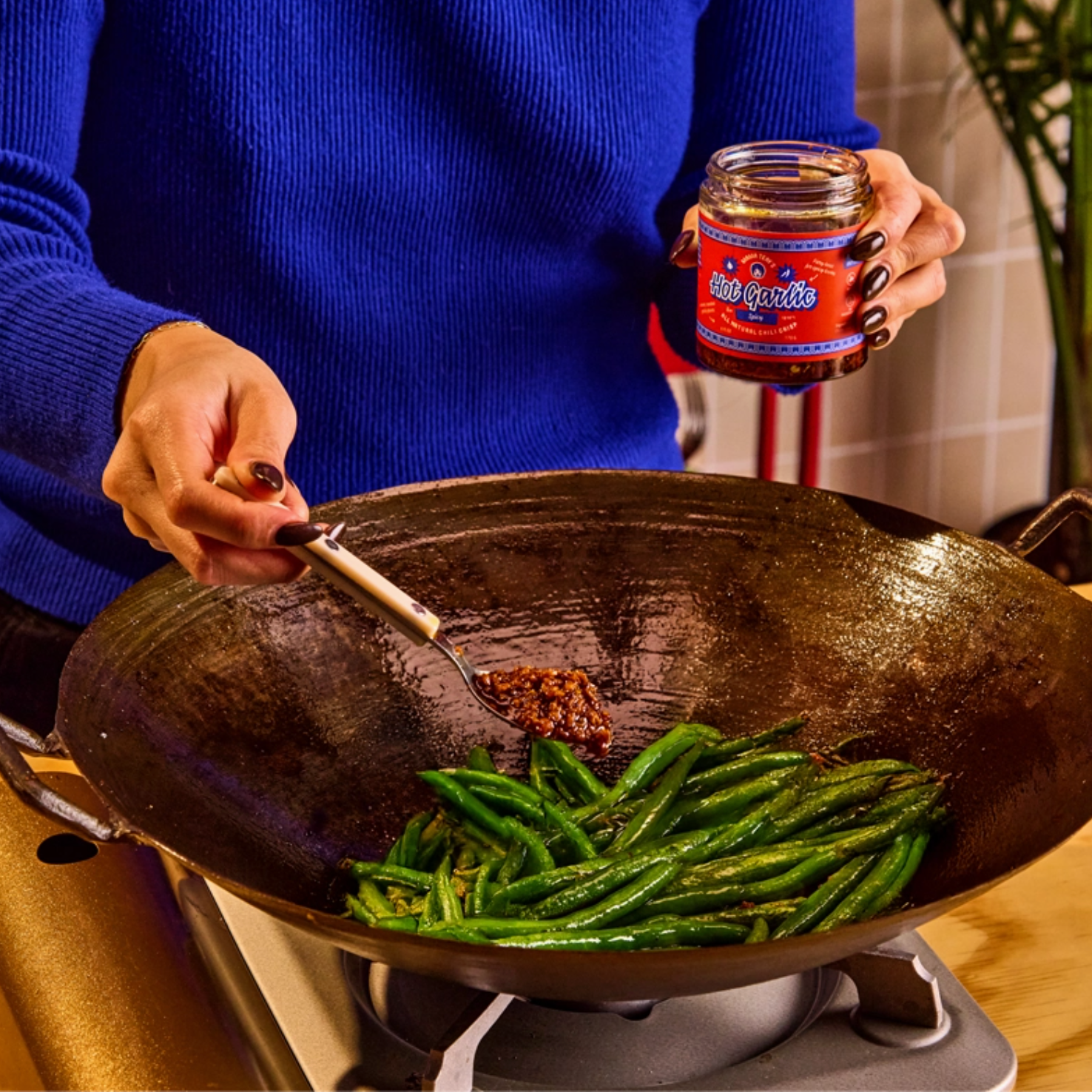 A person in a blue sweater adds a spoonful of chili oil from a jar labeled 'Red Hot Chili Sauce' to green beans cooking in a large black wok over a stove.