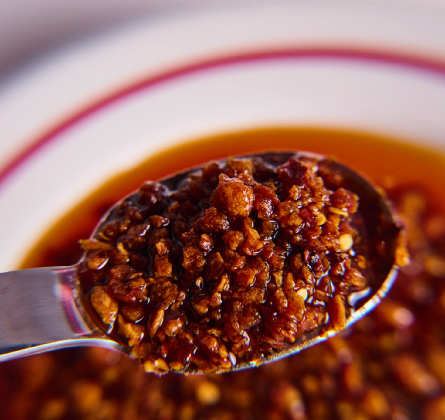 A close-up of a metal spoon filled with a glossy, chunky dark brown sauce or chili oil, set against a blurred white bowl with a red stripe.