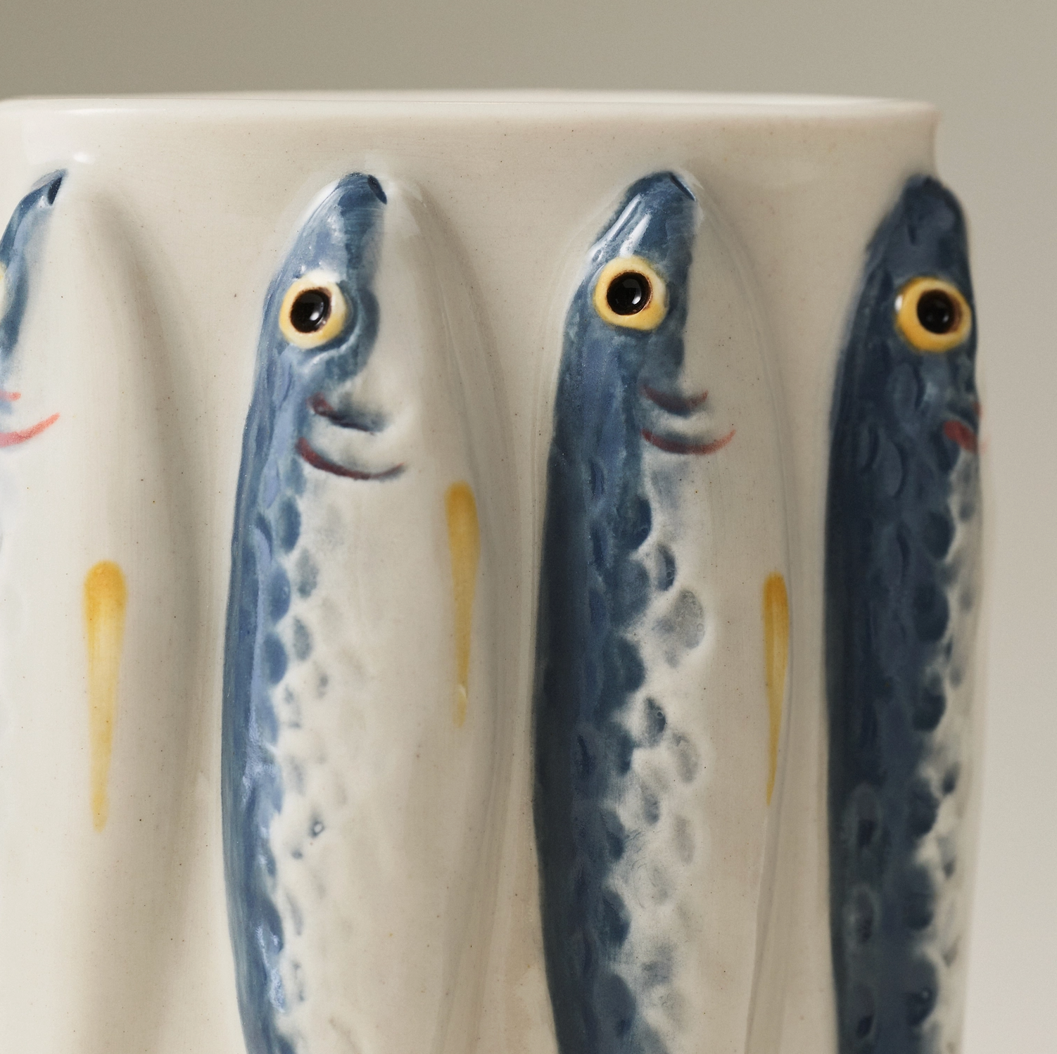 Close-up of a ceramic vessel with sculpted fish figures in blue, white, and yellow on a light neutral background.