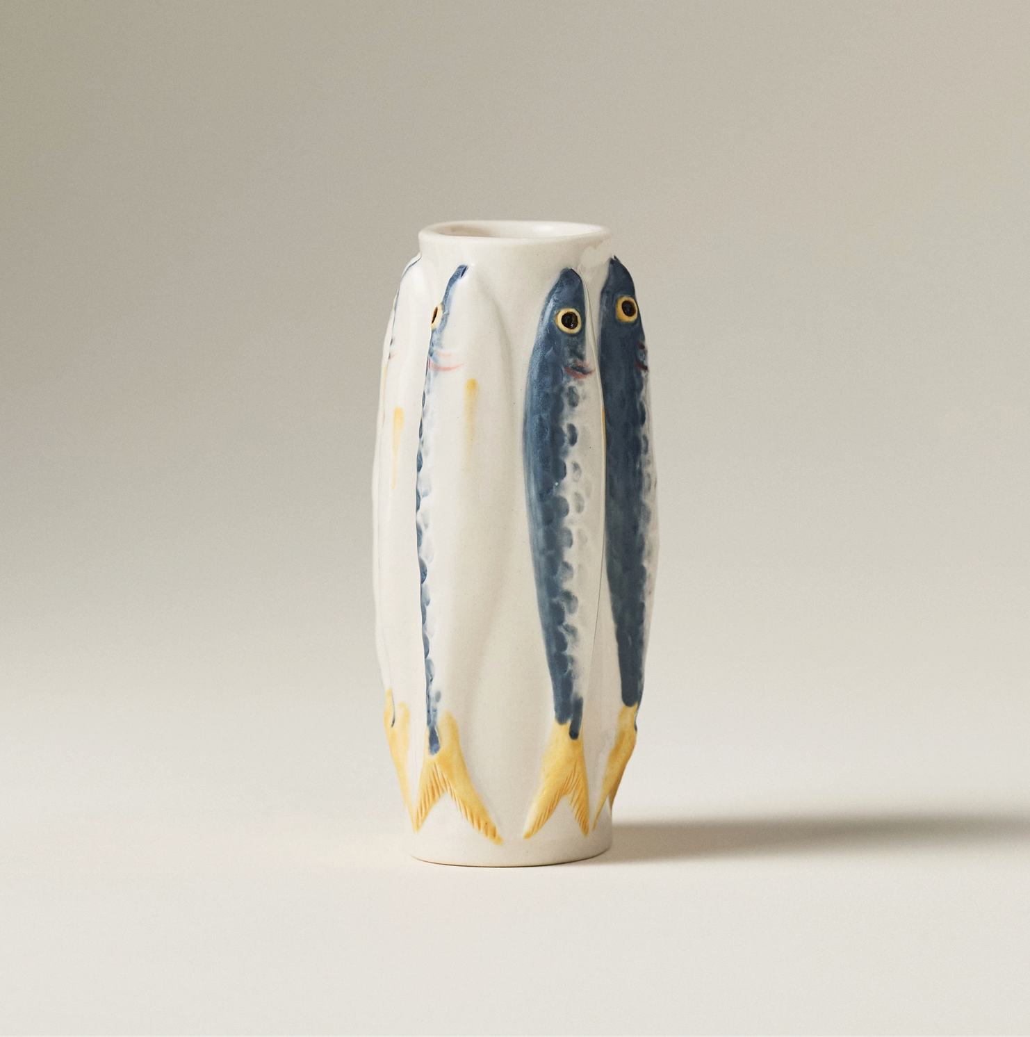 A white ceramic vase featuring three painted blue fish with yellow fins and gold eyes, set against a neutral light gray background.