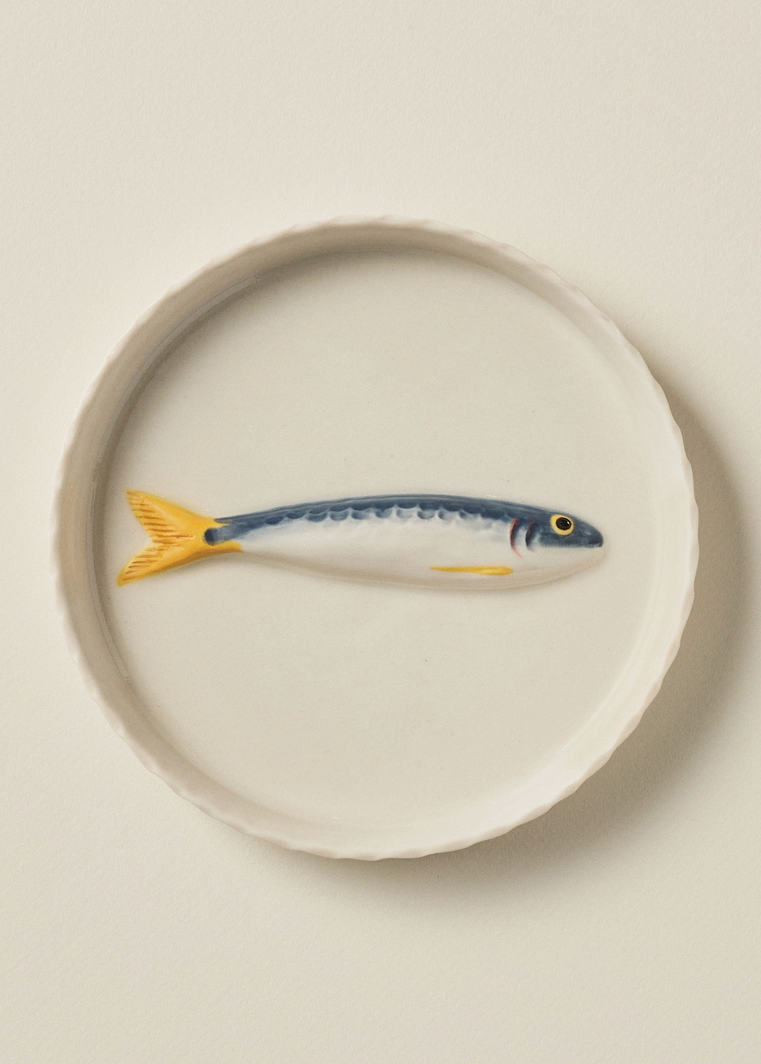 A round white ceramic plate featuring a painted blue and yellow fish design in the center, set against a solid light beige background.