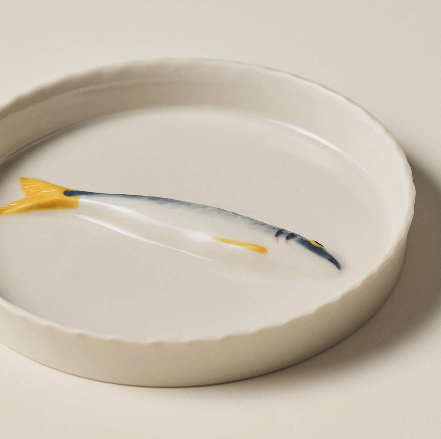 A white ceramic shallow bowl or dish with a scalloped rim, featuring a painted fish design in blue, yellow, and black on the inside surface, photographed against a warm off-white background.