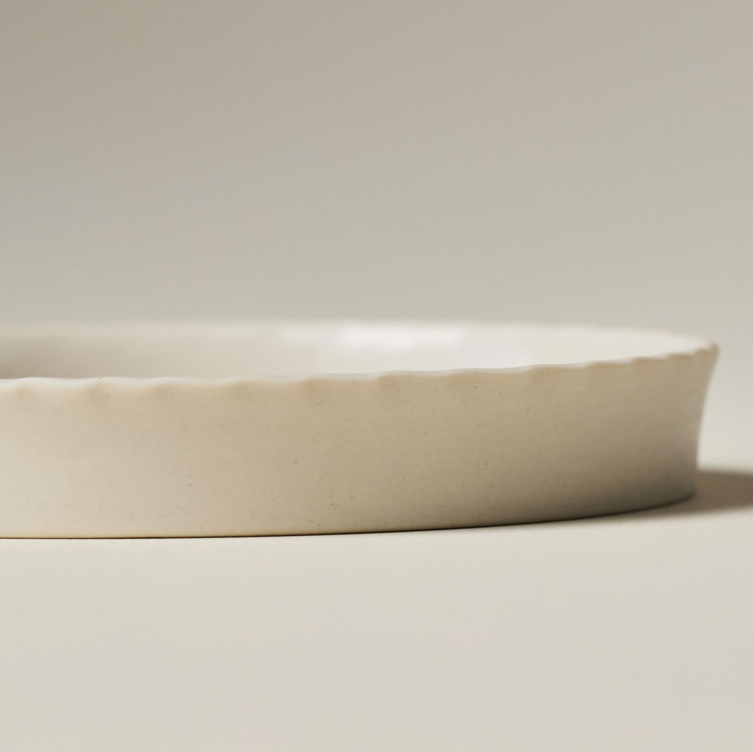 A close-up of a shallow round ceramic dish with a scalloped rim, shown in a warm off-white or cream glaze against a neutral light gray background.