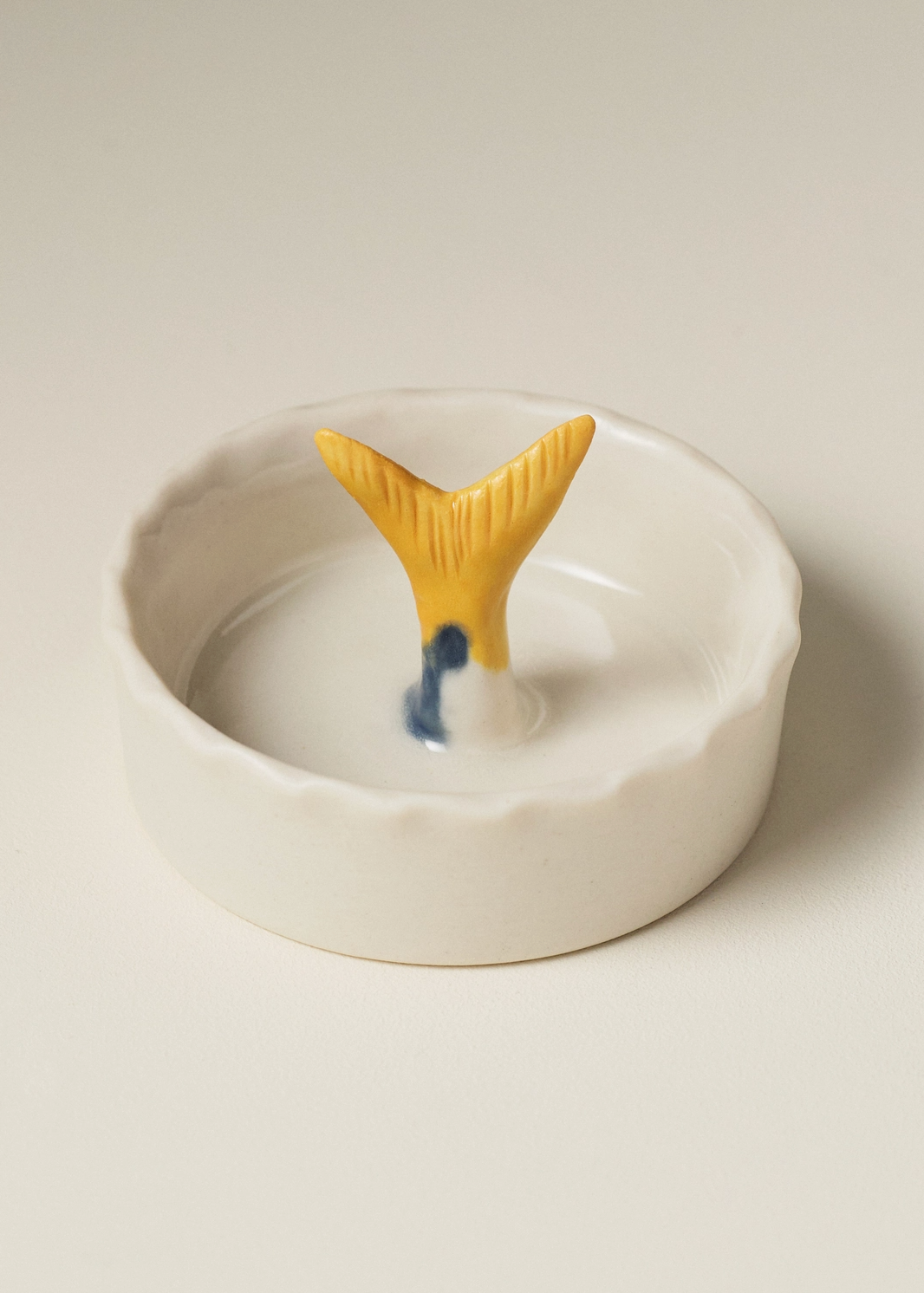 A small white ceramic dish with a scalloped rim and a yellow fish tail handle in the center, set against a plain light beige background.