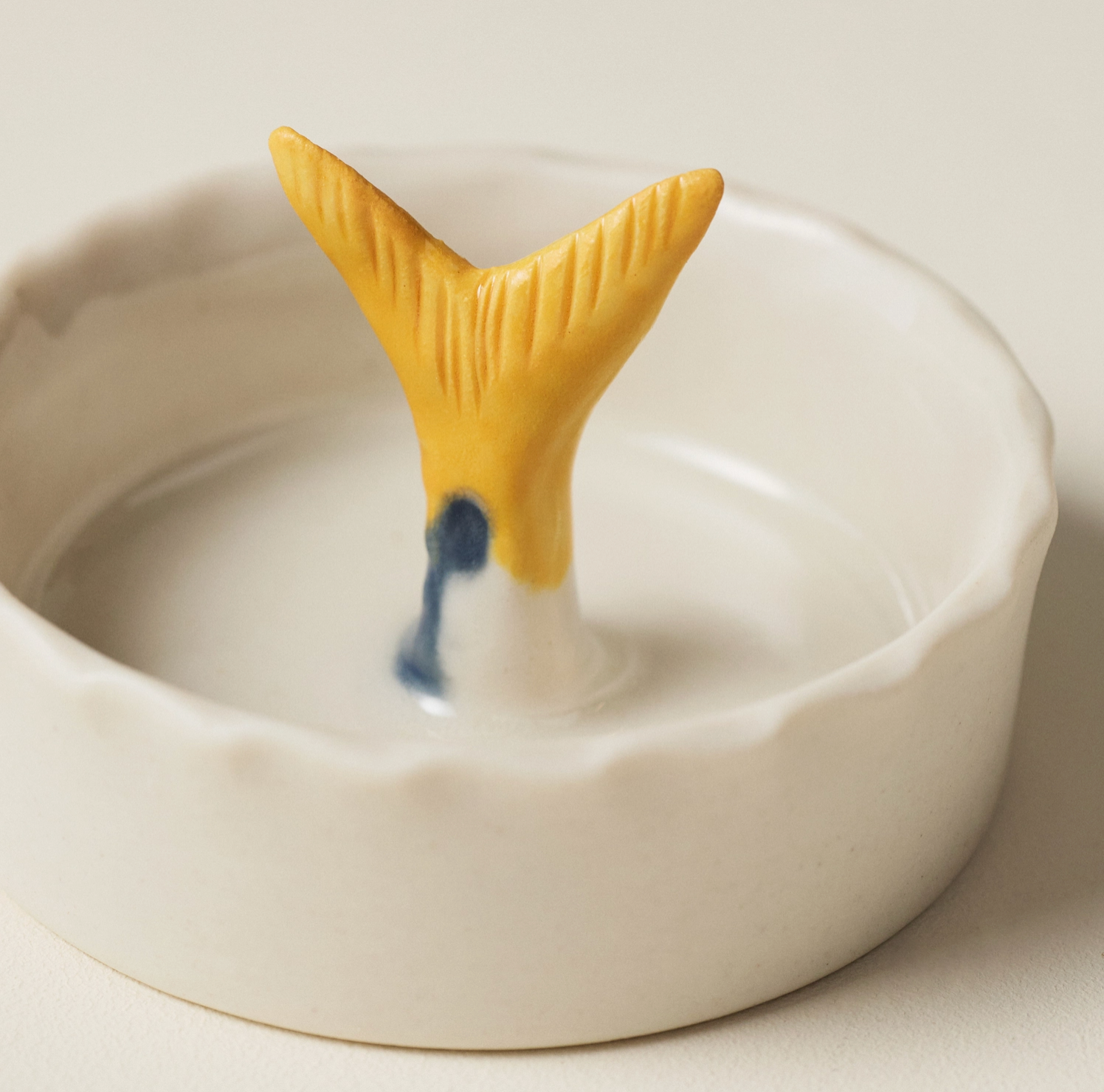 A white ceramic dish with a scalloped rim and a small yellow fish tail handle rising from the center, with a small blue accent near the base, photographed on a light neutral background.