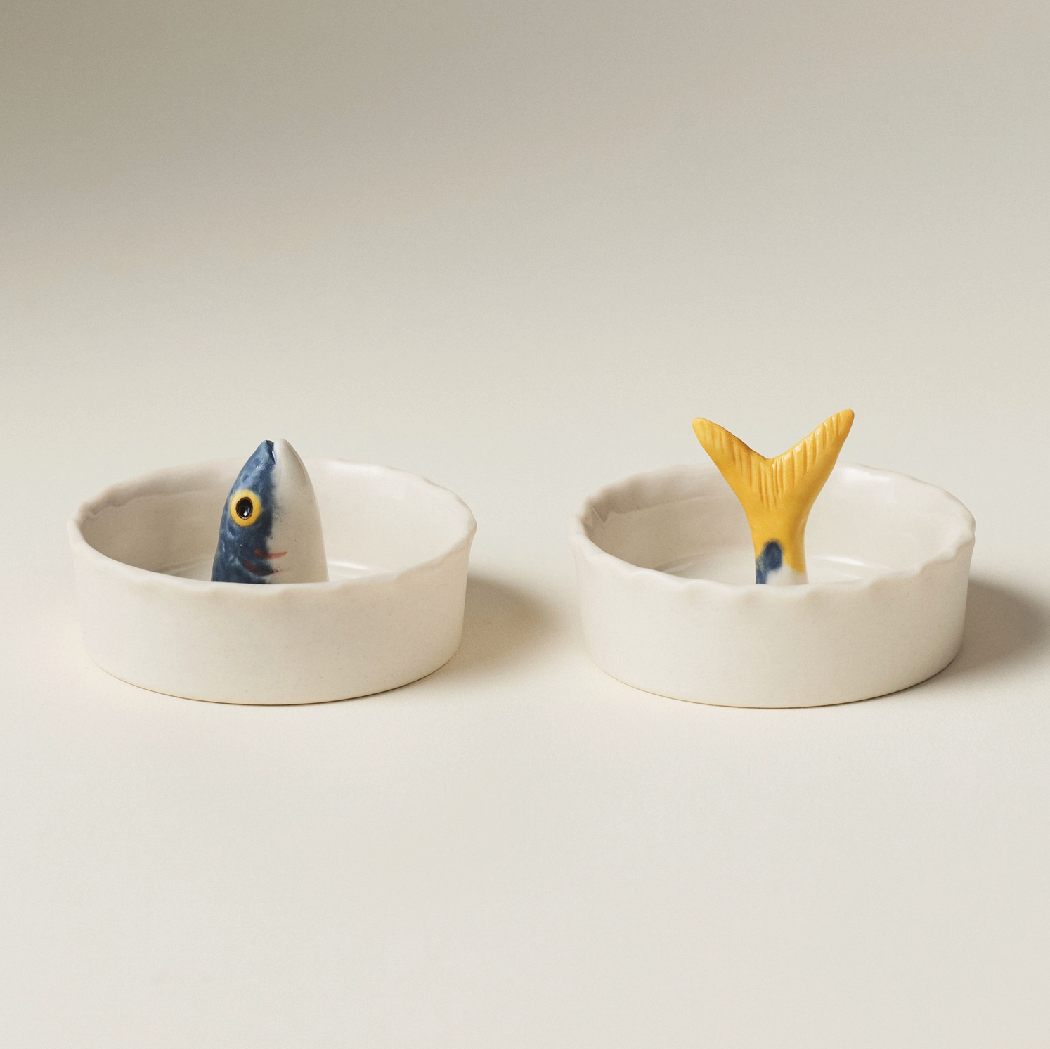 Two small white ceramic dishes with fish-shaped lids, one showing a blue and white fish head and the other showing a yellow fish tail, on a light beige background.