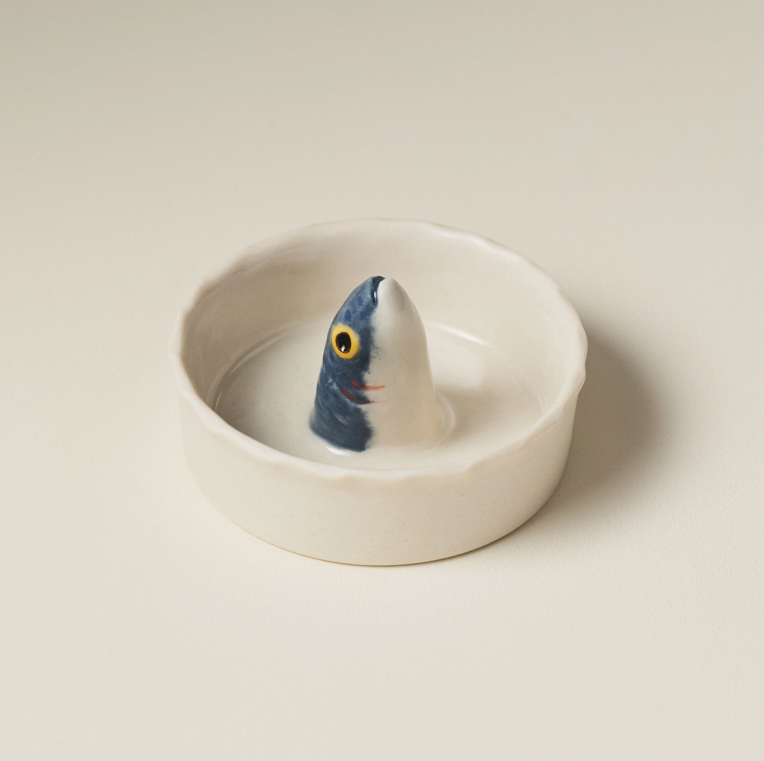 A small white ceramic dish with a sculpted fish-shaped insert, featuring a blue-gray body, white belly, and a yellow eye, photographed on a plain warm off-white background.