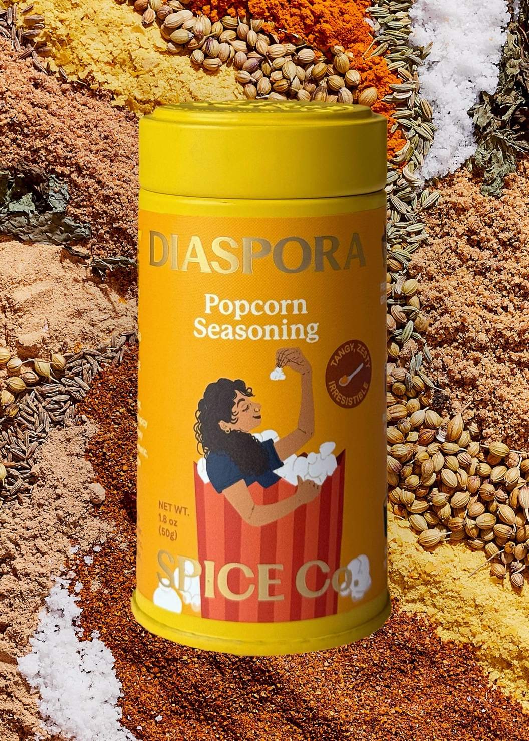 A yellow cylindrical canister of DIASPORA Popcorn Seasoning from Spice Co, set against a background of various spices, seeds, and herbs.