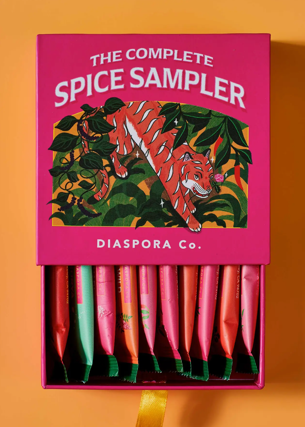 The Complete Spice Sampler box by Diaspora Co. shown open against a warm orange background, with a pink lid featuring a tiger illustration and multiple colorful spice sticks visible inside.