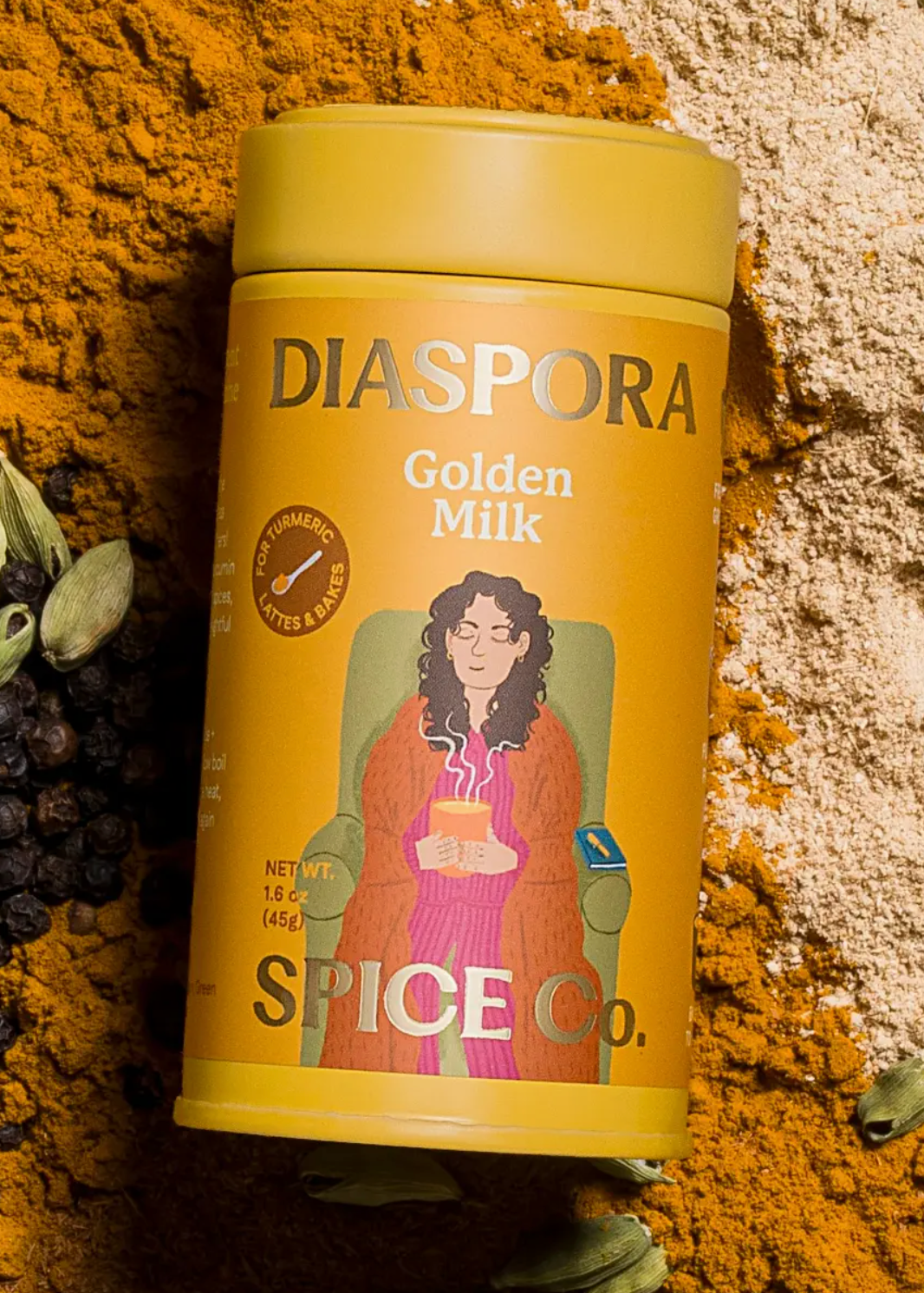 A yellow cylindrical tin of Diaspora Golden Milk Spice Co. is centered on a styled background of spices, including cardamom pods, black peppercorns, and a light beige powder.