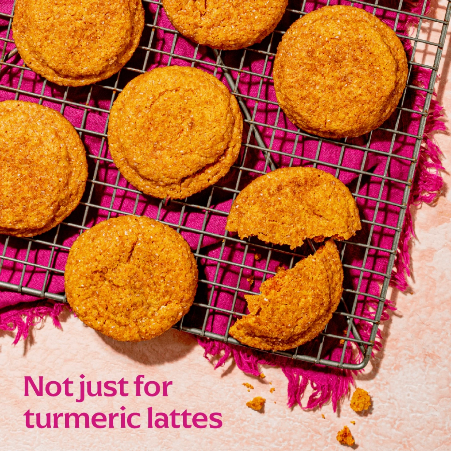 Several golden-brown cookies are arranged on a metal cooling rack over a bright pink textured cloth. One cookie is broken in half, and pink text in the lower left reads 'Not just for turmeric lattes'.