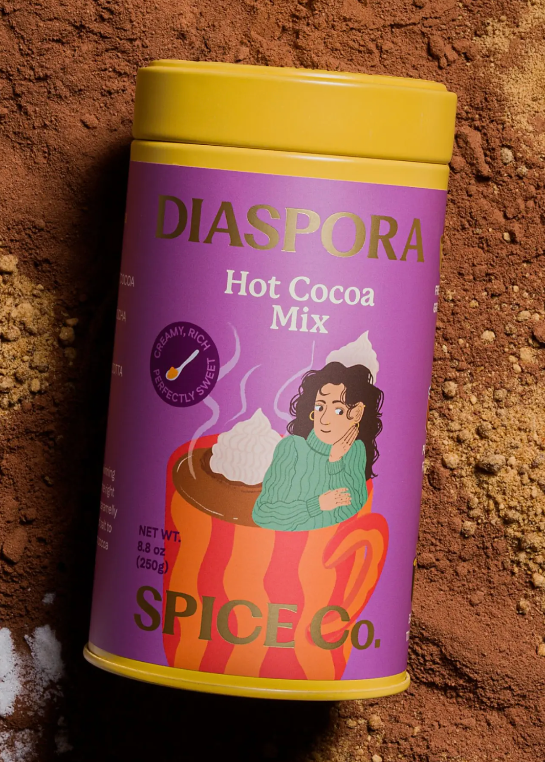 A tub of Diaspora Hot Cocoa Mix by Spice Co. stands upright on a textured brown powder background, with a small amount of white salt in the lower left corner.