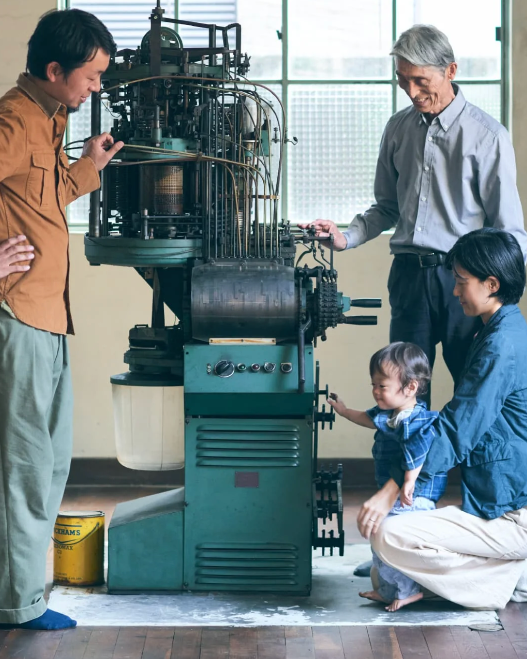 A family of four stands and crouches around a large vintage green textile spinning or knitting machine in a bright indoor workshop. An older man in a gray shirt stands behind the machine, while another man in a brown shirt points at it, and a woman and child look at the machine and its operation.