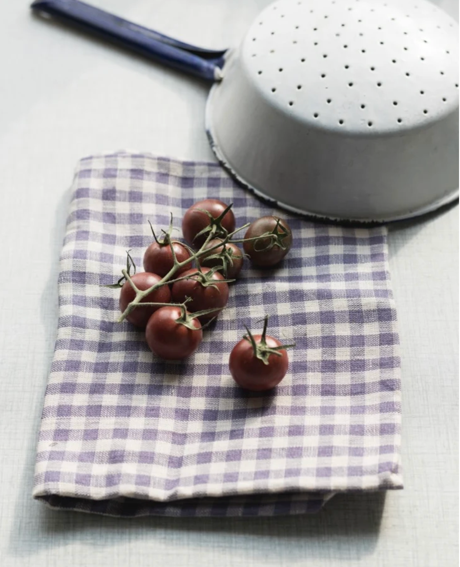 A folded checkered cloth napkin in lavender and white is styled on a light gray tablecloth, with a bunch of dark red cherry tomatoes on the napkin and the bottom edge of a white enamel colander visible in the background.