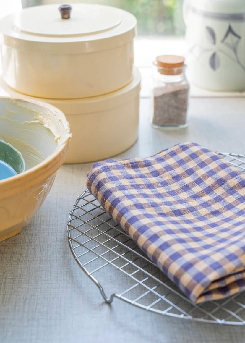A folded gingham kitchen towel rests on a metal cooling rack on a light gray tablecloth, with cream-colored ceramic kitchenware blurred in the background.