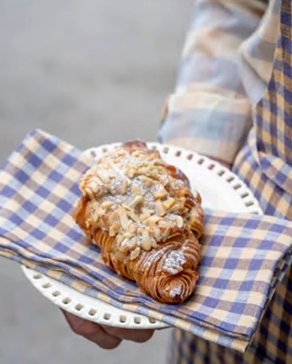 A close-up of a flaky croissant topped with sliced almonds and powdered sugar, served on a white plate with a blue-and-beige checkered napkin, held by a person wearing a matching plaid apron.