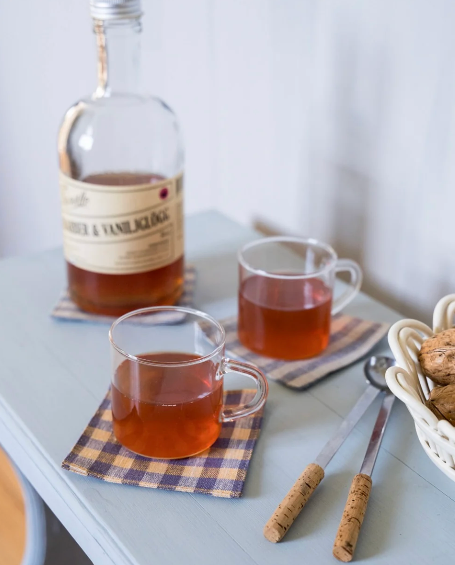 A clear glass bottle of whiskey and a pair of clear glass mugs filled with an amber drink, arranged on a pale blue-gray tabletop with checkered napkins, wooden-handled spoons, and a small basket of cookies.