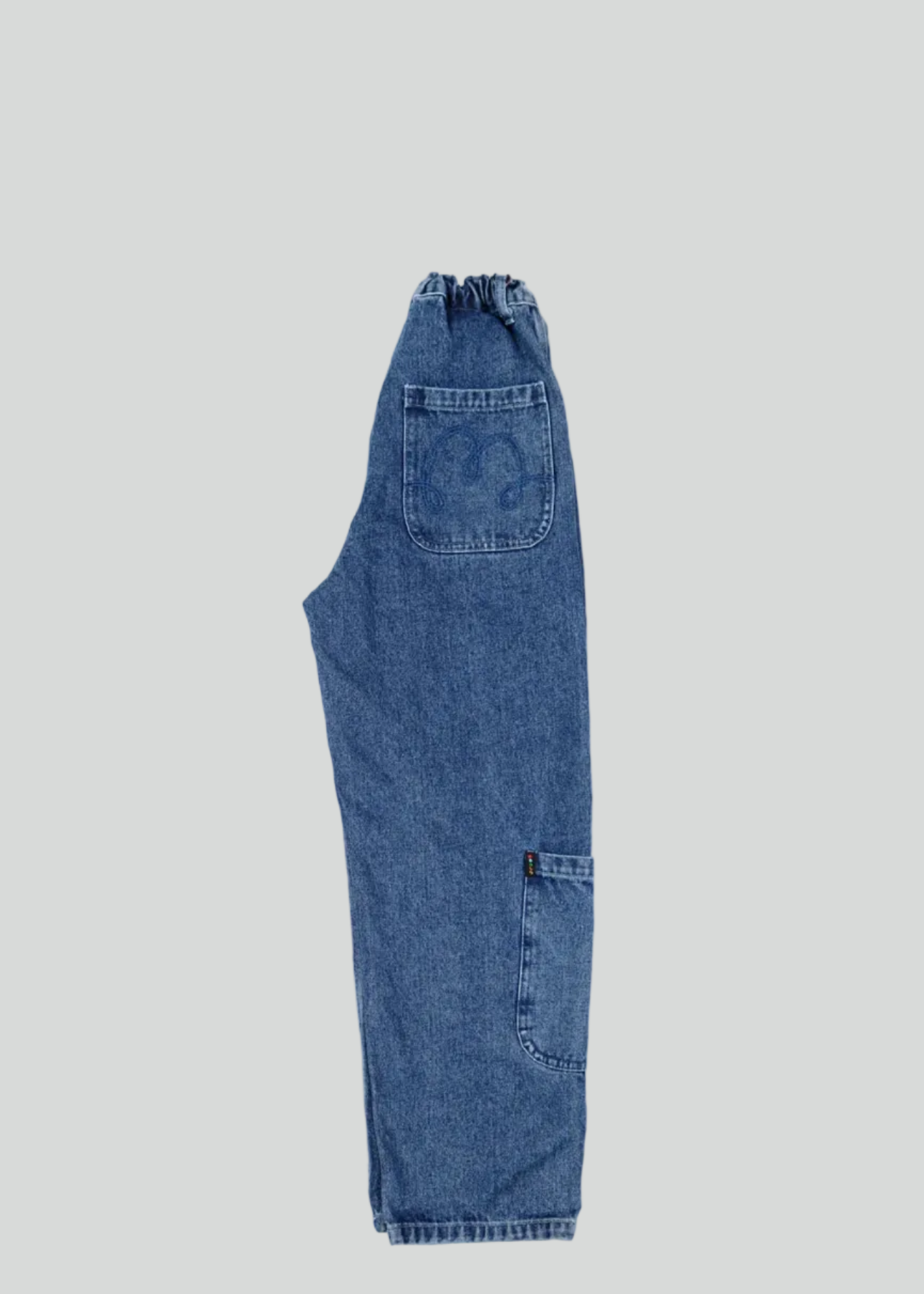 A pair of blue denim jeans shown flat against a white background, viewed from the back with a single large patch pocket and a cargo-style pocket on the lower left leg.