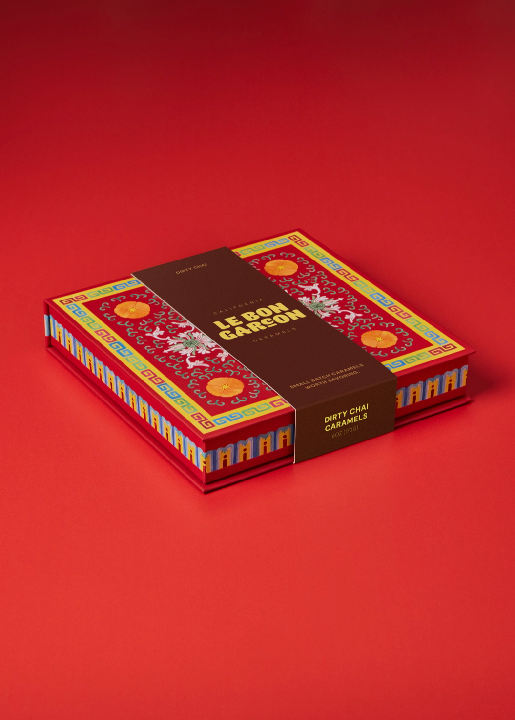 A boxed set of Le Bon Garcon Dirty Chai Caramels on a solid red background. The packaging features a red base with intricate gold, yellow, and blue patterns, wrapped in a dark brown paper band with the brand name 'LE BON Garcon" in gold lettering.