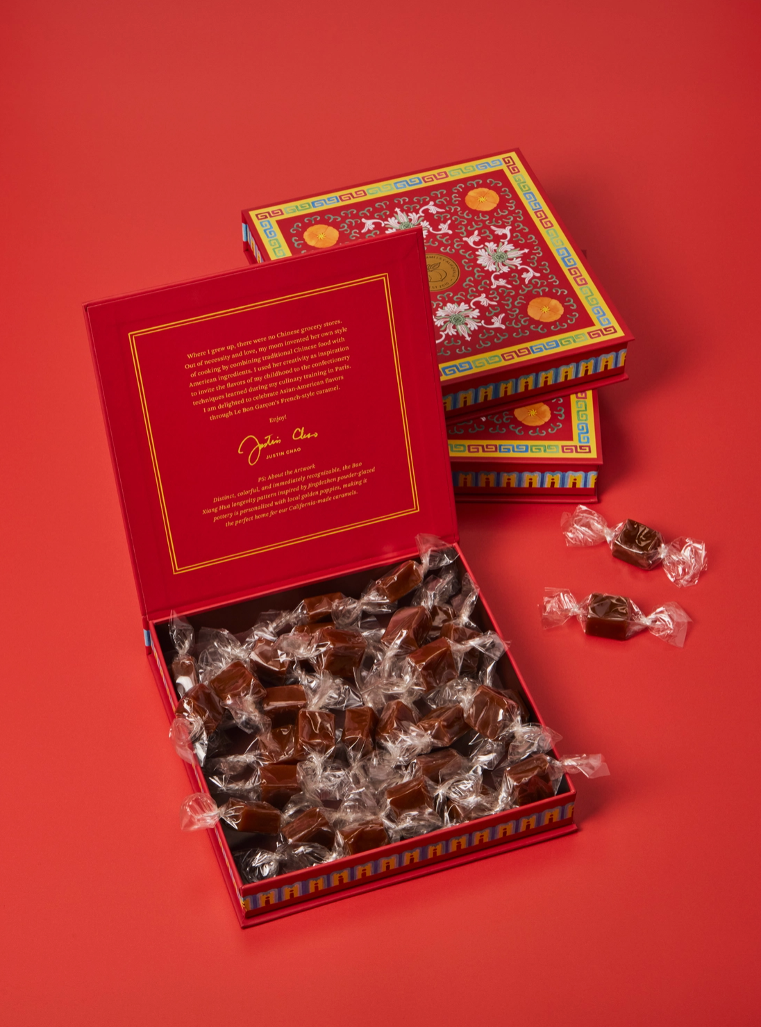 An open red gift box filled with individually wrapped caramels, with two closed decorative boxes stacked behind it on a solid red background. The boxes have ornate multicolor patterns and gold detailing, and the inside lid shows printed text and a signature.
