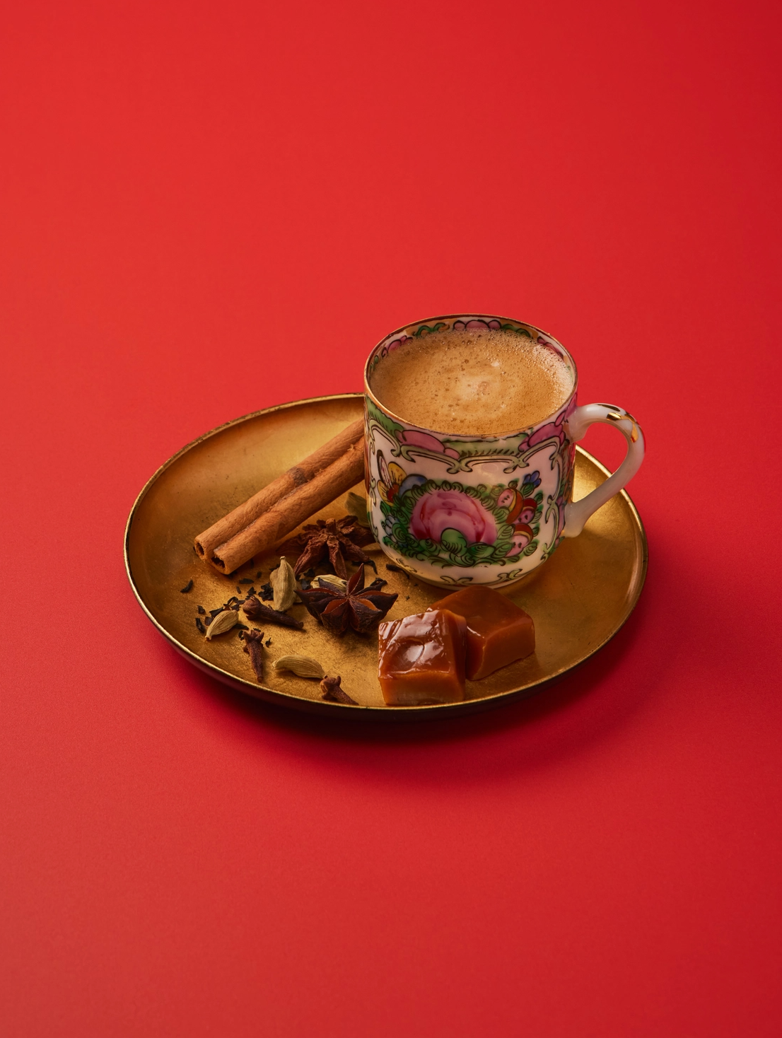A hand-painted ceramic Chinese coffee cup filled with coffee sits on a gold-toned metal tray, surrounded by cinnamon sticks, star anise, and caramel candies, all set against a solid red background.