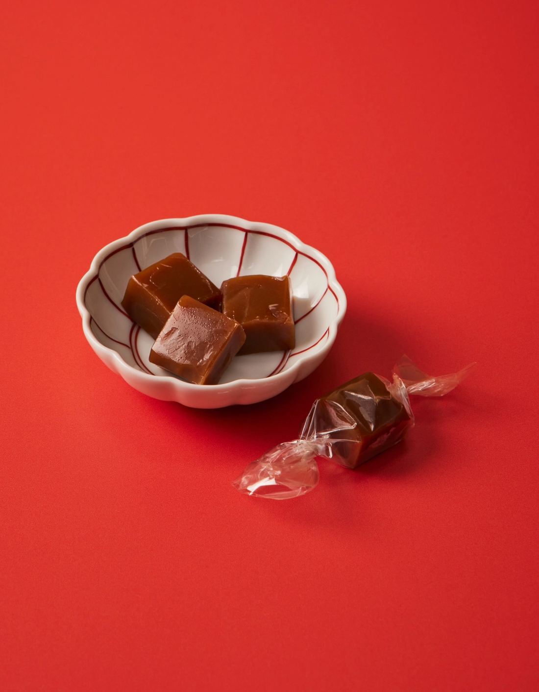 Three pieces of caramel candy in a small white ceramic bowl with red lines, next to one unwrapped caramel candy on a solid red background.