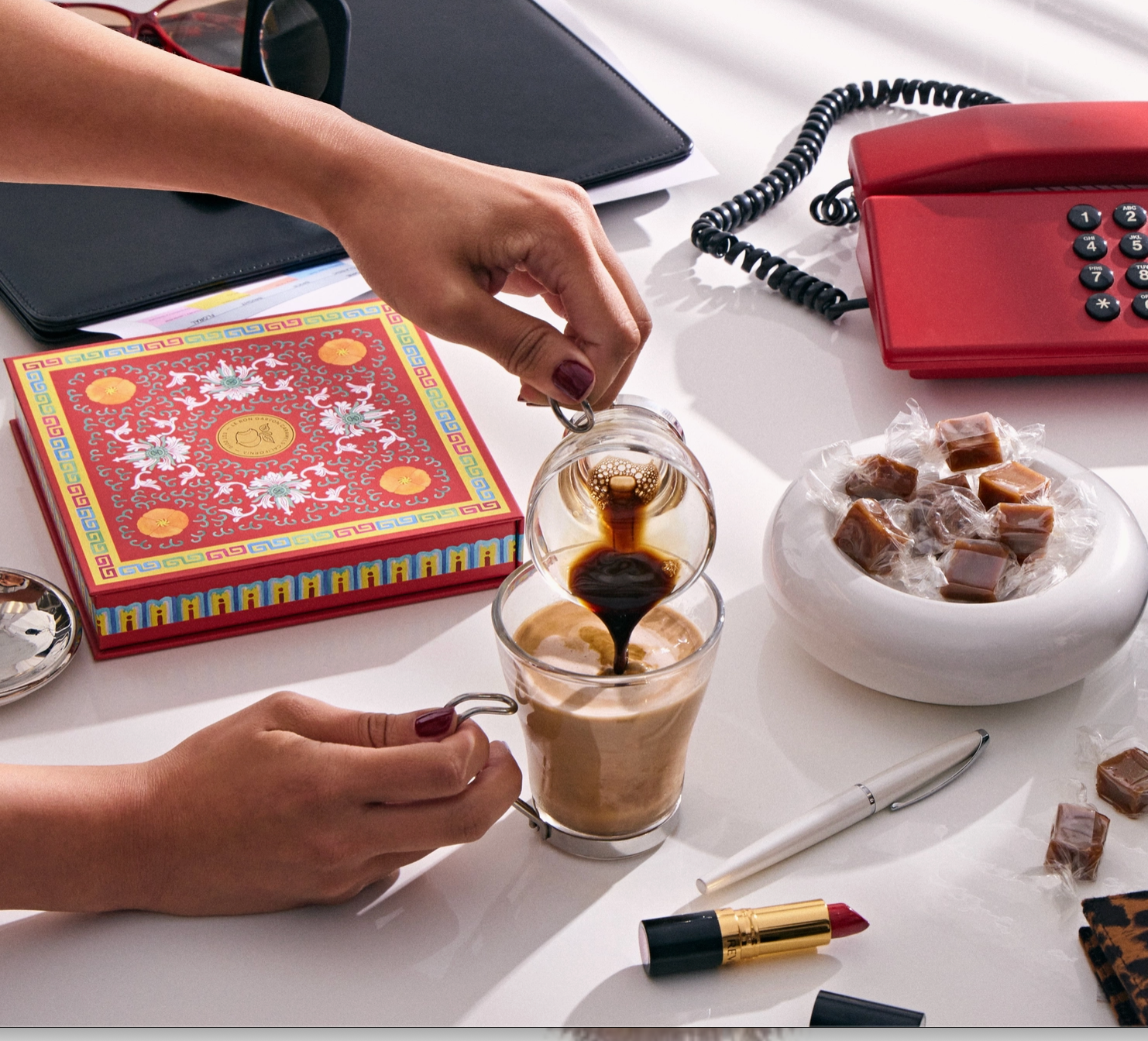 A lifestyle scene on a white table showing hands pouring dark coffee into a glass mug containing a light brown drink. Surrounding props include a red telephone, a decorative red box, a bowl of wrapped candies, lipstick, a pen, and a black folder.