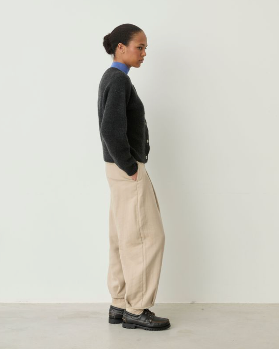 A model is shown in profile wearing a dark charcoal knit cardigan over a blue shirt, paired with beige wide-leg trousers and black leather shoes, standing against a plain off-white wall.