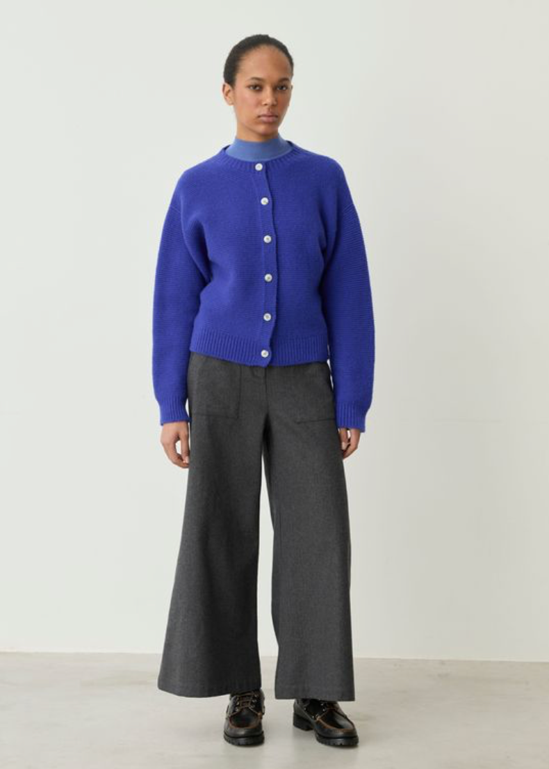 A model stands against a plain off-white studio wall wearing a bright royal blue button-front cardigan over a light blue mock-neck top, paired with dark gray wide-leg trousers and black leather shoes.