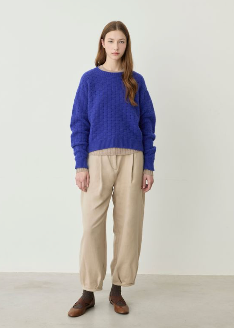 A woman stands against a plain off-white studio wall wearing a textured royal blue knit sweater, beige pleated trousers, and brown loafers.