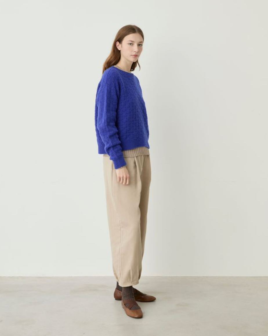 A woman stands in profile wearing a textured royal blue knit sweater, beige wide-leg knit pants, and brown leather shoes against a plain off-white studio background.