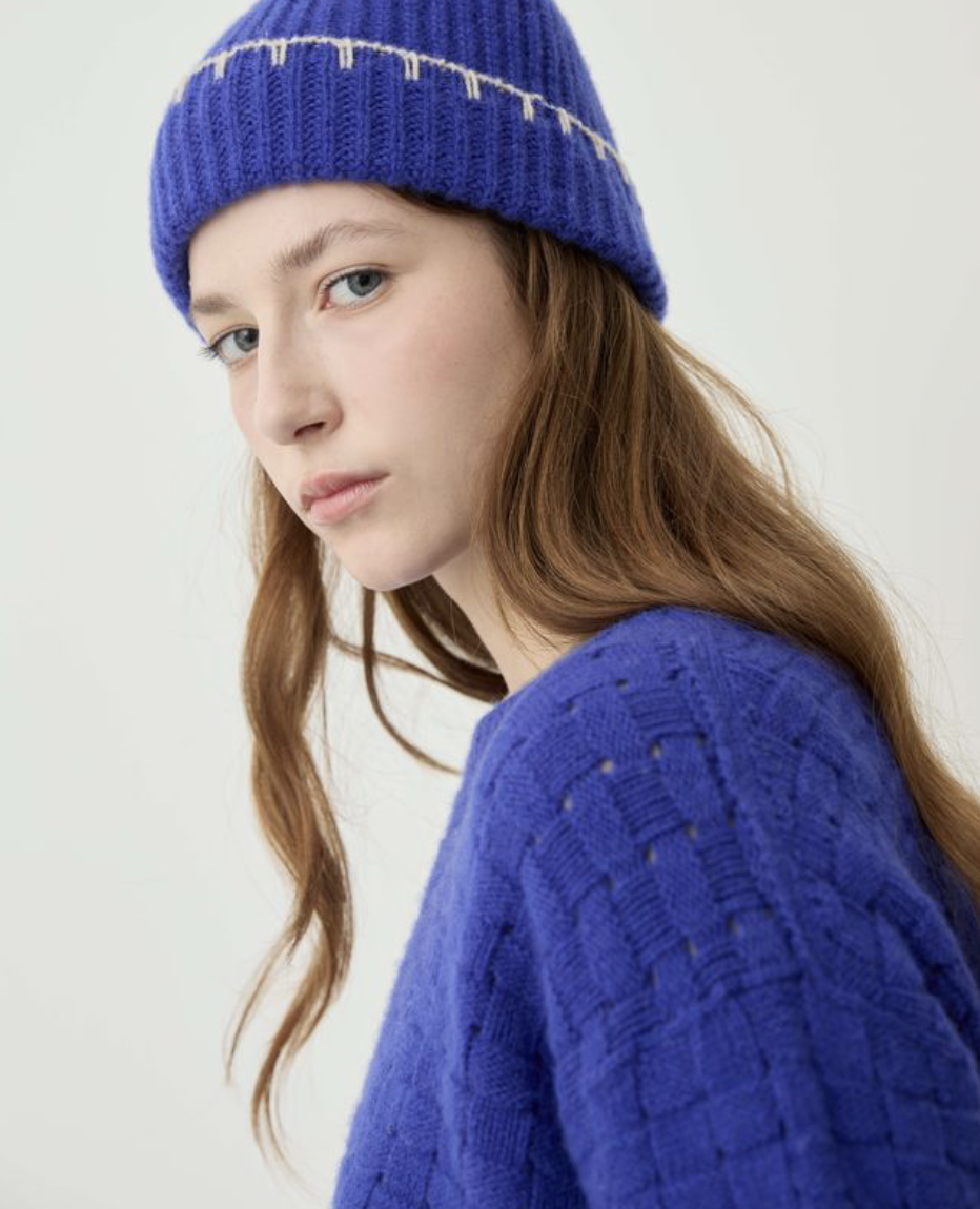 A woman wearing a bright blue knit beanie with a white zigzag stitch detail and a matching blue textured knit sweater, photographed against a plain light background.