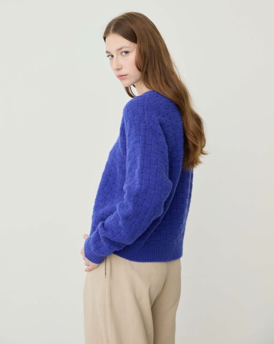 A woman wearing a textured royal blue knit sweater and beige trousers, photographed against a plain off-white studio background.