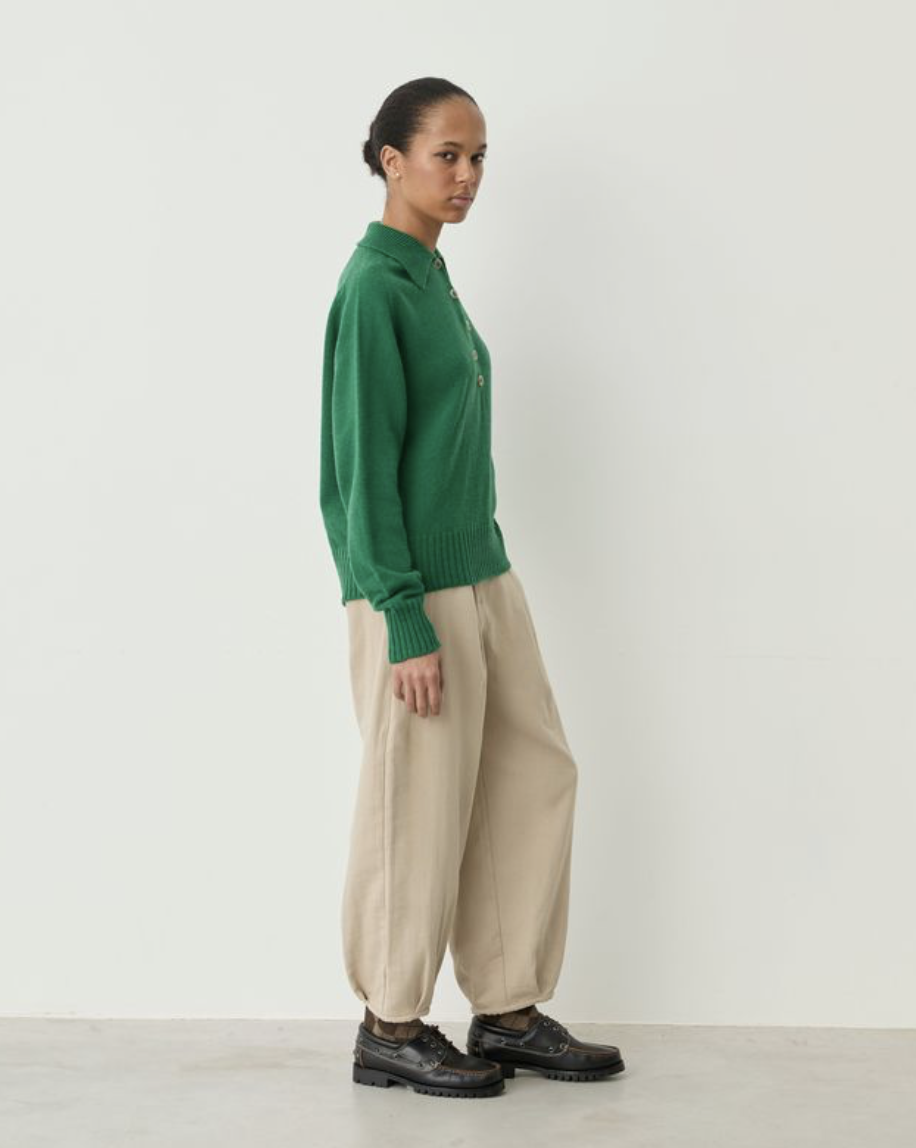 A model stands in profile wearing a green knit sweater with gold buttons, beige wide-leg trousers, and black leather shoes against a plain off-white studio wall.