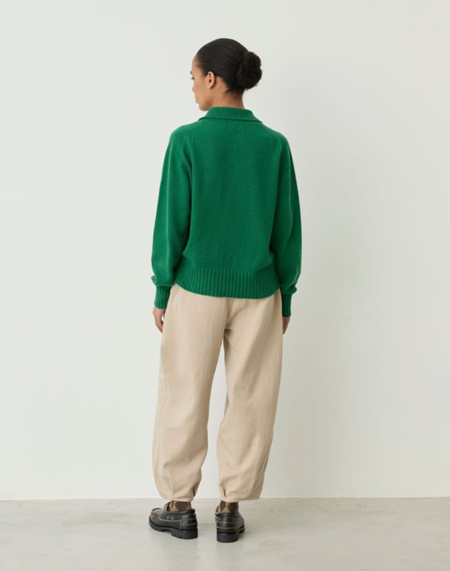 A model shown from the back wearing a vibrant green knit sweater, beige wide-leg trousers, and black leather shoes, standing against a plain off-white wall.