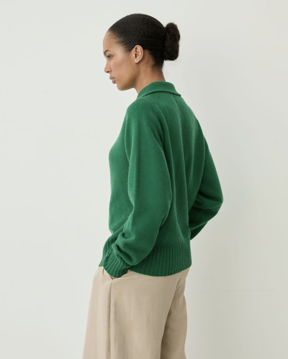 A woman shown from the side profile wearing a green knit sweater and beige trousers, standing against a plain light gray studio background.