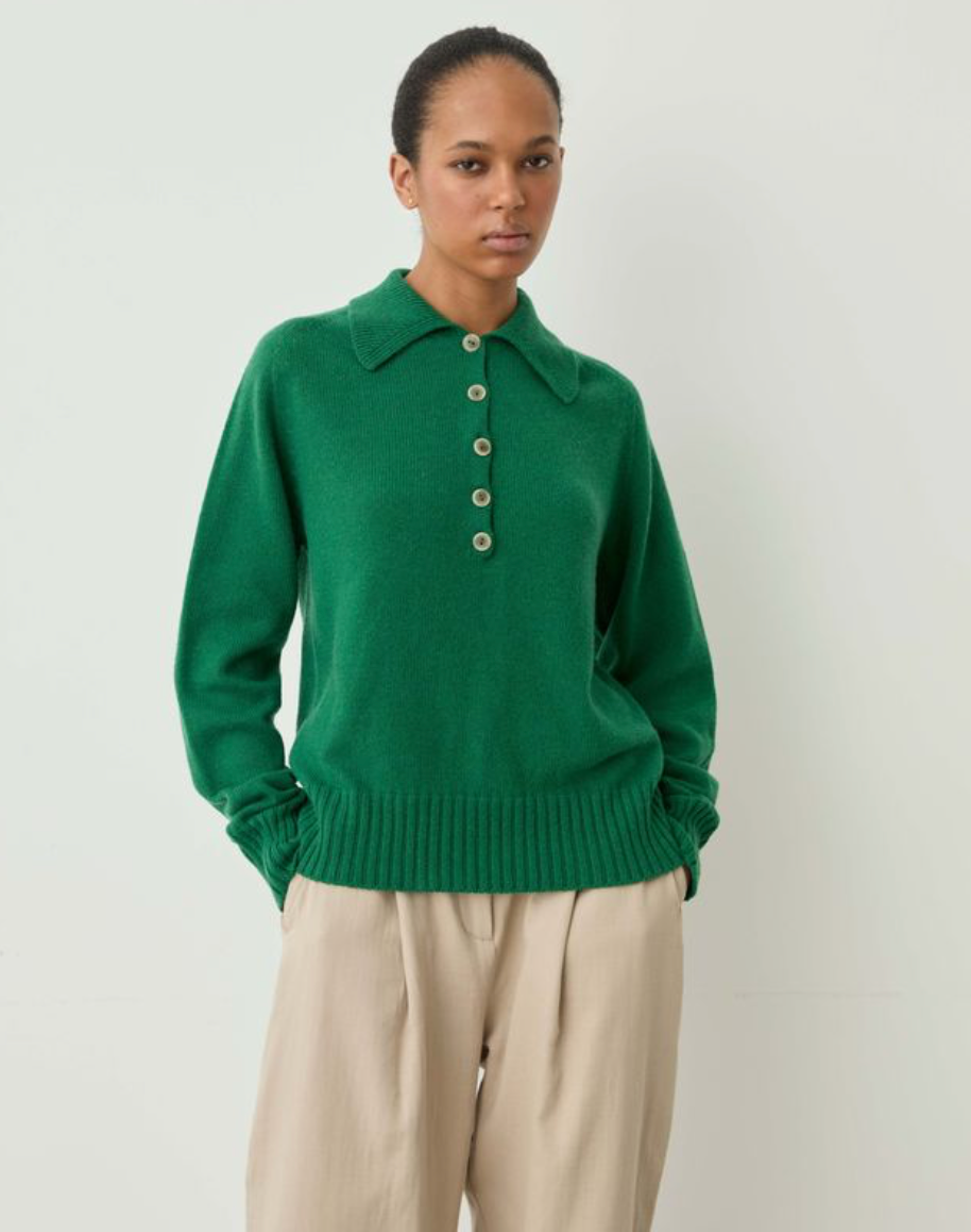 A model wearing a vibrant green knit polo sweater with a buttoned placket and beige trousers, standing against a plain off-white studio background.