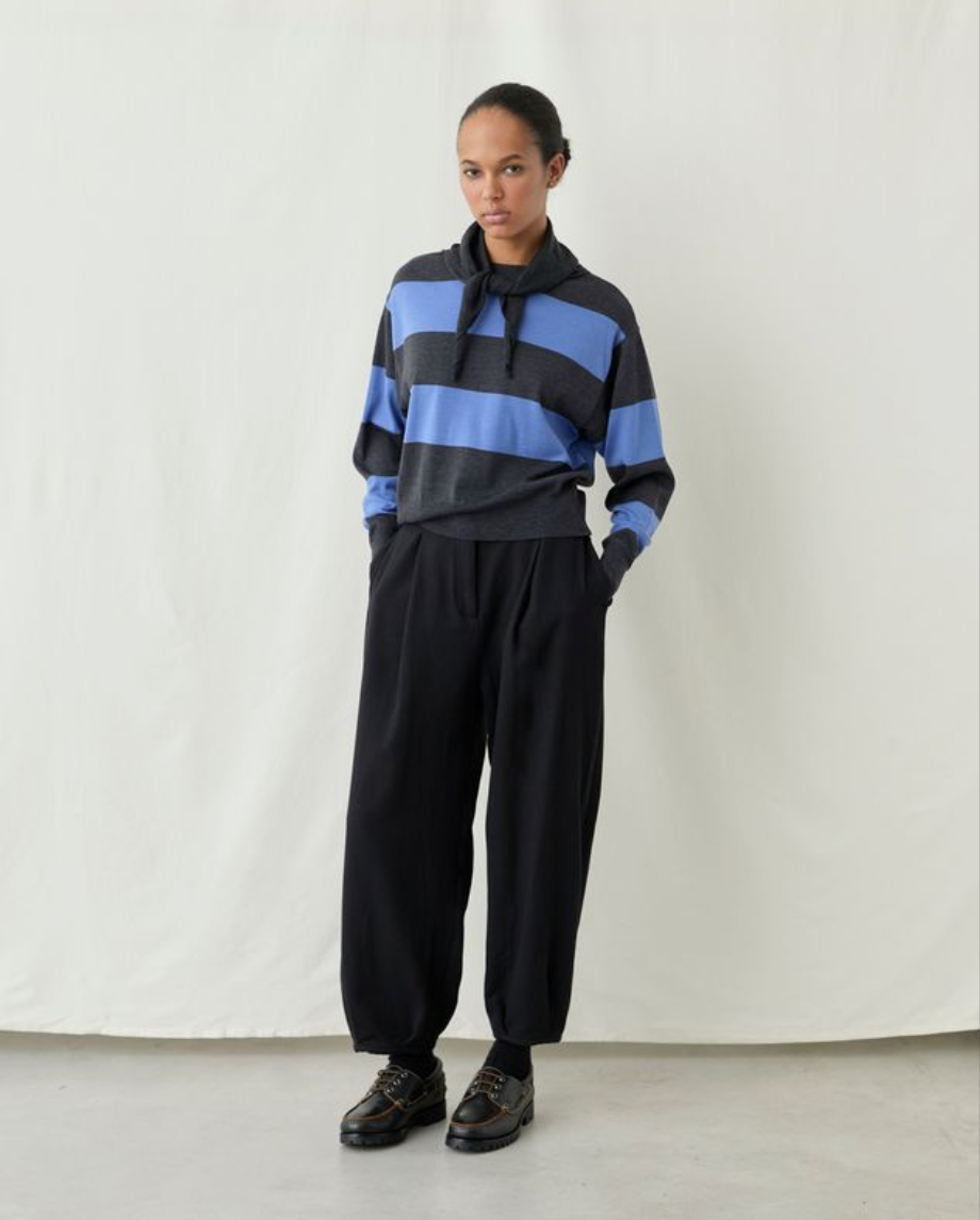 A model stands against a white studio backdrop wearing a dark gray and blue horizontally striped hoodie with black drawstrings, paired with loose black sweatpants and dark brown leather shoes.