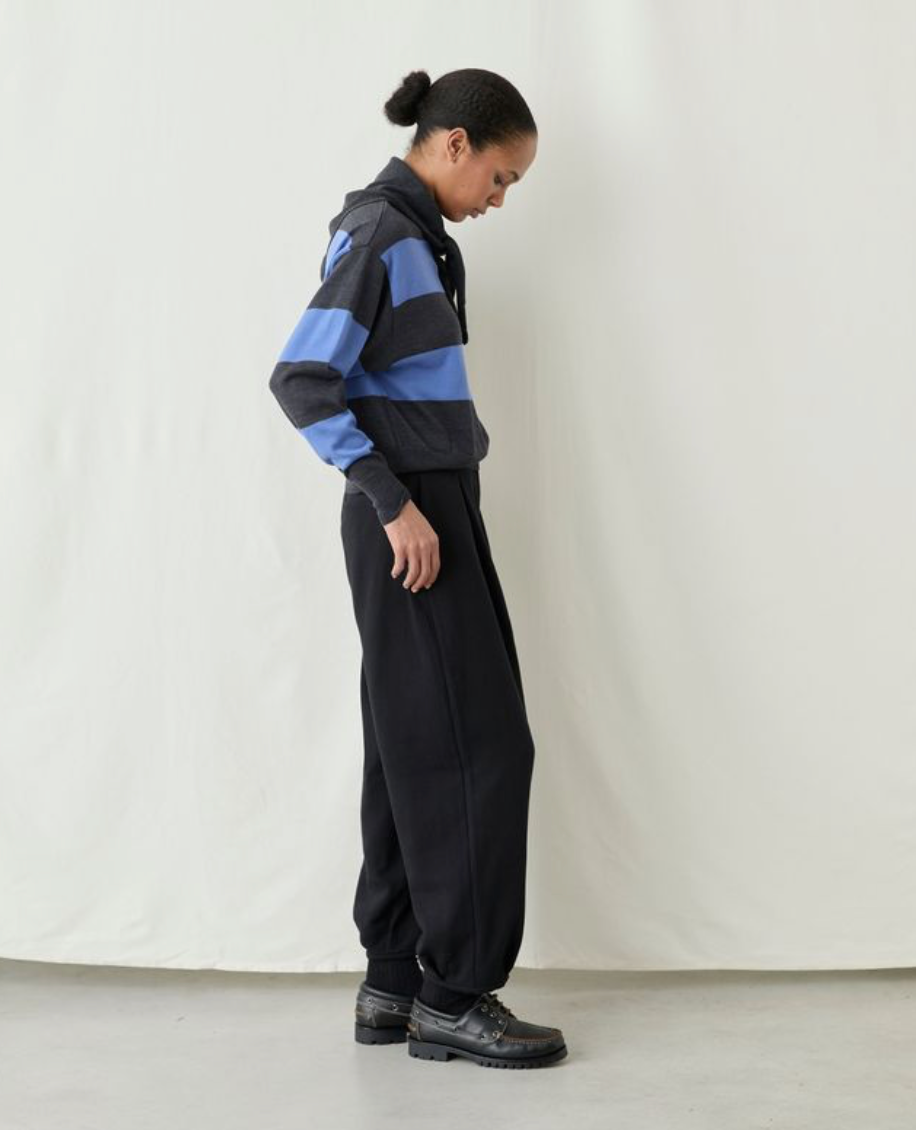 A model wearing a black and blue striped hoodie with black sweatpants and black leather shoes, standing in profile against a white studio backdrop.