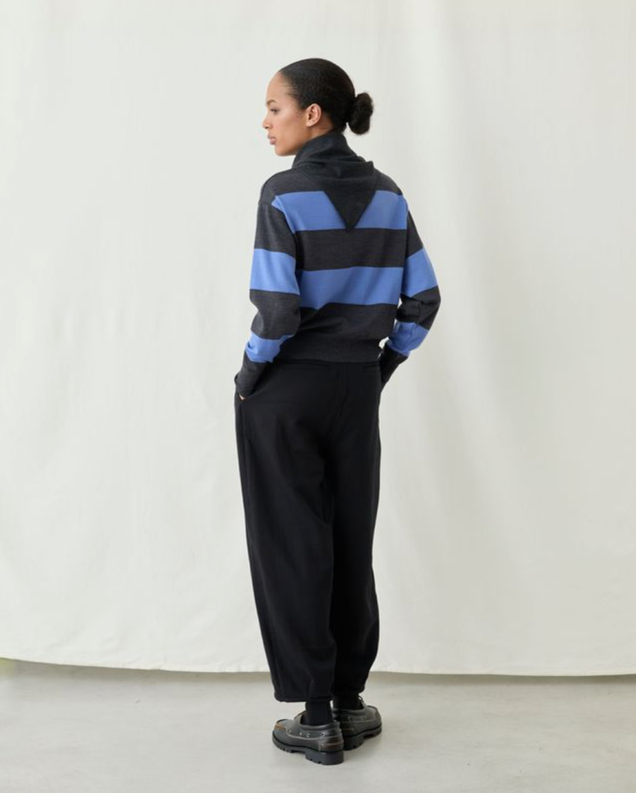 A model stands in profile wearing a black and blue striped mock-neck knit sweater and black wide-leg trousers against a plain off-white studio backdrop.