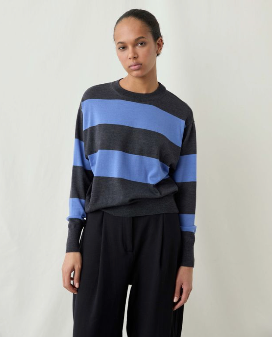 A model wearing a dark gray and light blue striped crewneck sweater with black wide-leg trousers, standing against a plain off-white studio background.