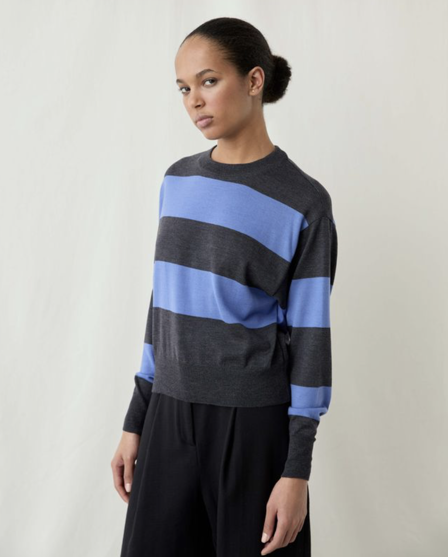 A model wearing a dark gray and light blue striped knit sweater with black trousers, photographed against a neutral off-white studio background.