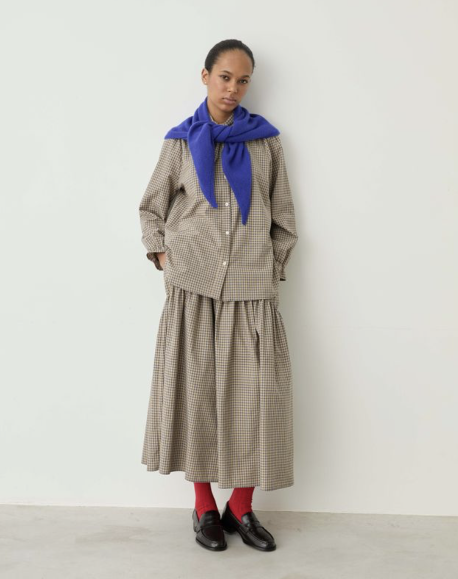 A model stands against a plain off-white wall wearing a beige and brown gingham button-up shirt and matching pleated midi skirt, styled with a vibrant royal blue scarf, red socks, and black leather shoes.