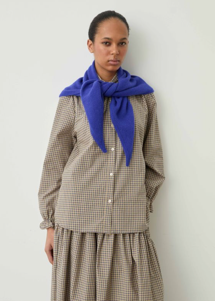 A model stands against a plain light gray wall wearing a brown and beige gingham long-sleeve shirt and matching skirt, with a bright royal blue knit scarf tied around the neck.