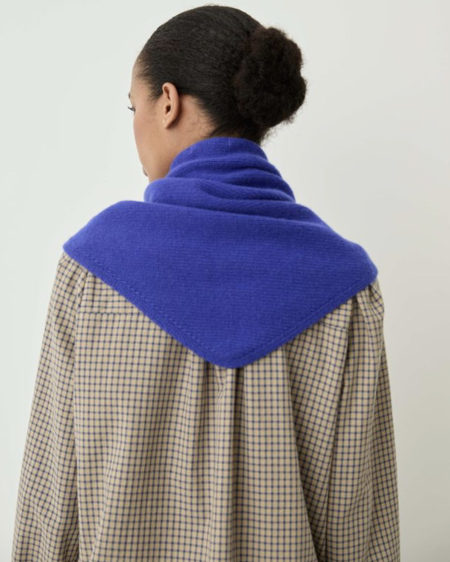 A person shown from the back wearing a bright royal blue knit neck warmer or cowl layered over a beige and black checkered shirt, against a plain light background.