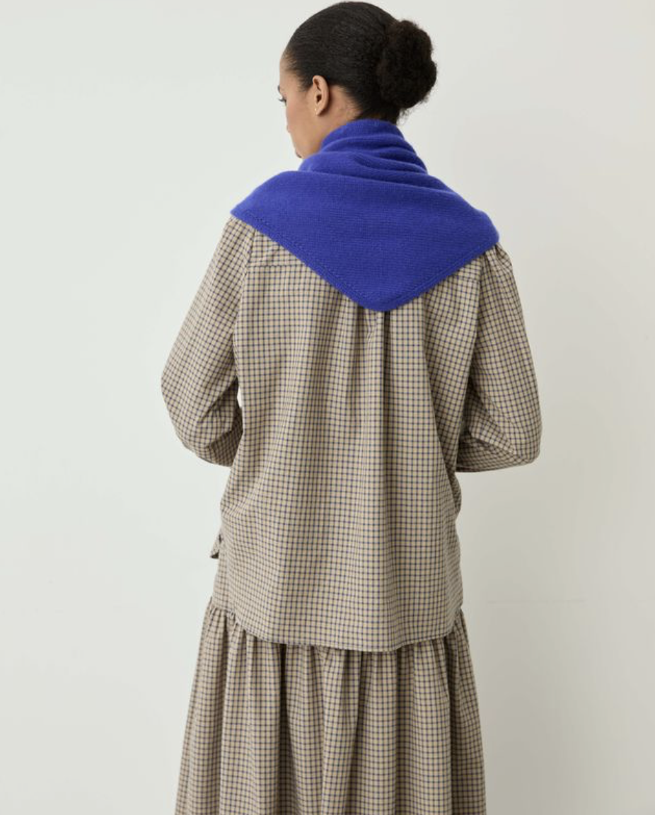 A woman shown from the back wearing a brown and beige gingham dress with a vibrant royal blue knit collar, set against a plain light gray background.