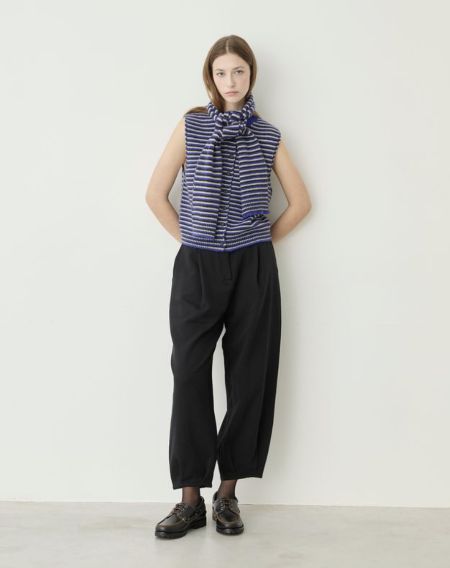 A model stands against a plain off-white wall wearing a sleeveless top with horizontal navy and white stripes and a large front bow, paired with loose black pants and dark lace-up shoes.
